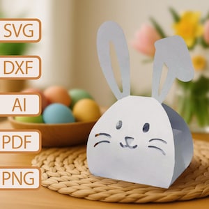 May include: A white paper bunny rabbit shaped box with cut-out ears, eyes, and nose. The box is sitting on a woven wicker surface.