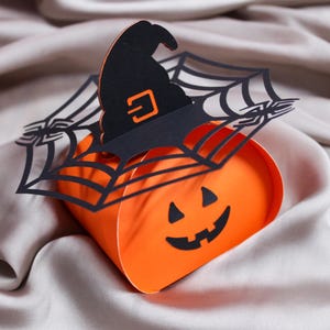 Può includere: Una scatola di carta arancione con una faccia di zucca nera, sormontata da un ritaglio di carta nera a forma di cappello da strega e ragnatela. L'immagine include etichette per i formati DXF, SVG, PDF, JPG, PNG e il marchio Lunavy.