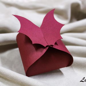 Może przedstawiać: Bordowa papierowa skrzynka na prezent z dekoracyjną górą. Pudełko ma kwadratowy kształt ze złożonym wzorem i stylizowaną ozdobą w kształcie liścia na górze. Tło to miękka, neutralna tkanina.