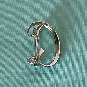 May include: Silver-toned letter D brooch with a decorative, stylised design. The letter is in a script font.