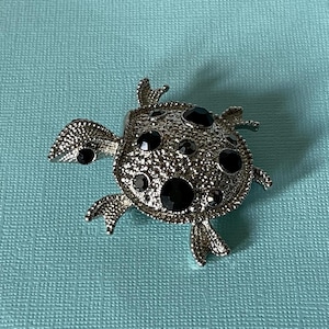 May include: A silver turtle brooch with black rhinestones. The turtle is detailed with a textured shell and has a shiny finish.