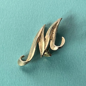 May include: A gold-colored metal brooch in the shape of the letter "M". The brooch has a stylized, cursive design with a textured surface. The pin is set against a light blue background.