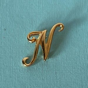 Może przedstawiać: Złota broszka w kształcie litery N. Litera jest w stylu pisma odręcznego.