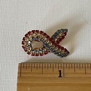 May include: A red, white, and blue jeweled ribbon pin. The pin is about 1 inch long.