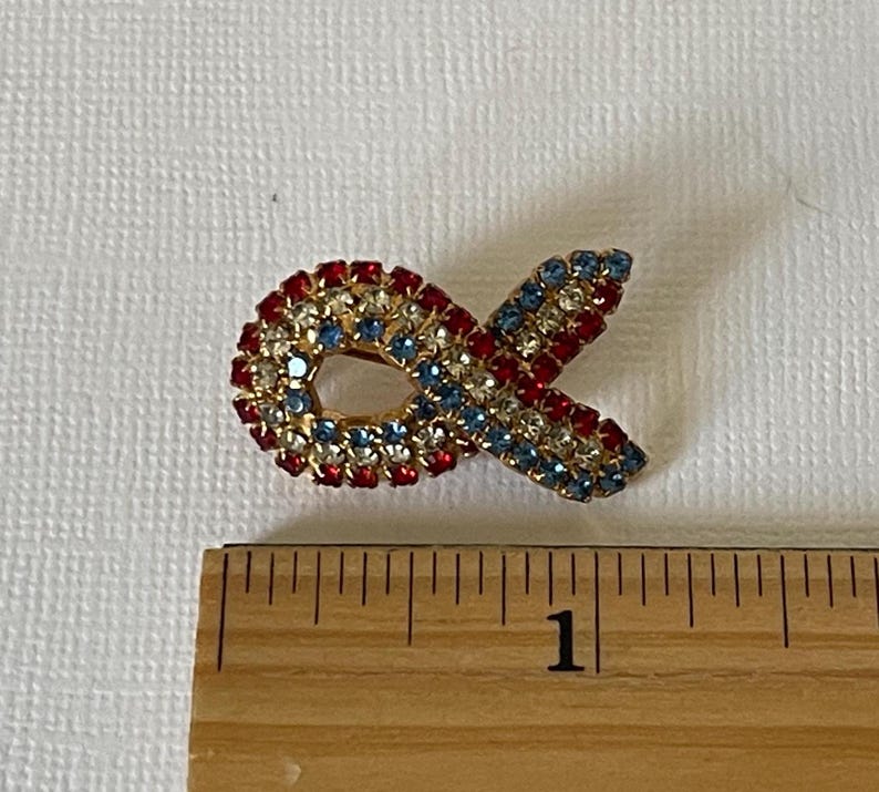 May include: A red, white, and blue jeweled ribbon pin. The pin is about 1 inch long.