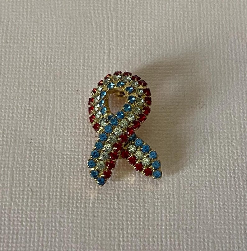 May include: A red, white, and blue jeweled ribbon pin. The ribbon is made of rhinestones and is in the shape of a bow.