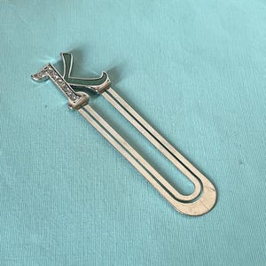 May include: Silver-toned metal bookmark with the letter "K" in a script font. The letter is embellished with clear rhinestones and a green enamel accent. The bookmark has two parallel, slender metal strips.