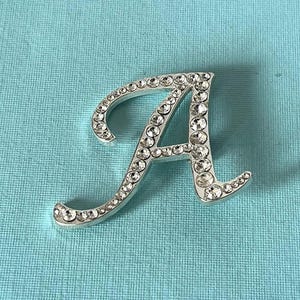 May include: Silver-toned letter "A" embellished with numerous clear rhinestones. The cursive font is set against a light blue textured background. This decorative initial is suitable for jewelry or craft projects.