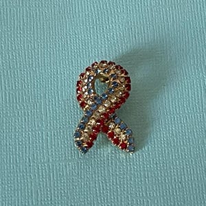 May include: A red, white, and blue jeweled ribbon pin. The ribbon is made of rhinestones and is in the shape of a bow.