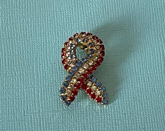 May include: A red, white and blue jeweled ribbon pin. The ribbon is made of rhinestones and is in the shape of a bow.
