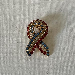 May include: A gold-toned pin shaped like a red, white, and blue ribbon, covered in rhinestones.