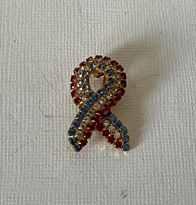 May include: A gold-toned pin shaped like a red, white, and blue ribbon, covered in rhinestones.