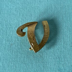 Peut inclure: Une épingle en métal doré en forme de la lettre "V". L'épingle a un dessus incurvé et un fond droit et ouvert. L'épingle est sur une surface texturée bleu clair.