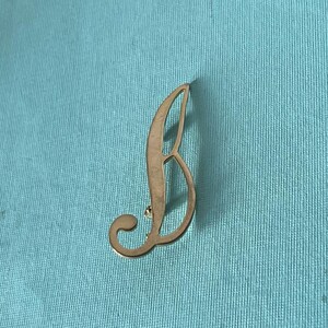 Peut inclure: Une épingle en métal doré en forme de lettre B. La lettre est dans un style d'écriture cursive.
