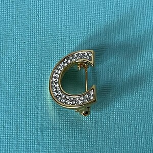 May include: A gold-toned letter C brooch with clear rhinestones. The brooch is on a turquoise background.