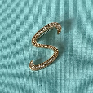 May include: A gold-toned brooch in the shape of the letter "S", embellished with small, clear crystals. The brooch is set against a light blue textured background, accentuating the jewellery's design.