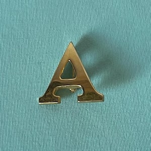 May include: A gold-colored letter A pin. The letter is cut out, creating a hollow center.