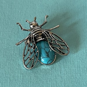 May include: A silver bee brooch with a turquoise stone in the center. The bee's wings are detailed and intricate.
