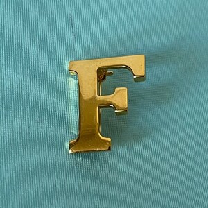 May include: A gold-colored letter F brooch. The letter is raised and has a shiny finish.