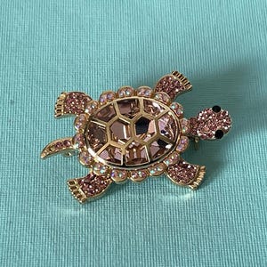 May include: A gold-toned turtle brooch with a shell of faceted pink and purple crystals. The turtle's head and flippers are adorned with smaller pink crystals. Black accents form the eyes. A decorative accessory.