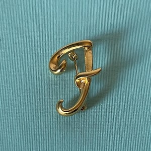 May include: A gold-toned letter F brooch with a cursive script design.