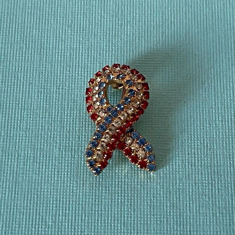 May include: A red, white, and blue jeweled ribbon pin. The ribbon is made of rhinestones and is in the shape of a bow.