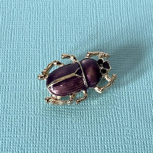 May include: A gold-toned beetle brooch featuring a deep purple enamel finish on its wings and body. The insect-shaped pin displays detailed legs and a segmented head, with a polished metallic gold border surrounding the vibrant purple enamel sections.