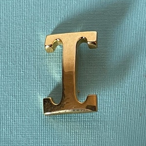 May include: A gold-colored letter "T" with a shiny, reflective surface. The capital letter is displayed against a textured, light blue background. The letter is a solid, three-dimensional object.
