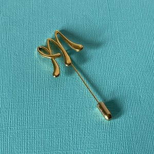 Może przedstawiać: Złota przypinka do krawata ze stylizowanym wzorem litery "M". Przypinka ma długą, smukłą igłę i ozdobną górę w kursywie. Przypinka jest umieszczona na turkusowym tle.