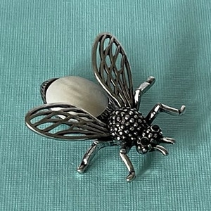 May include: A silver bee brooch with a white pearl-like body and black accents. The wings are detailed with a delicate wire design.