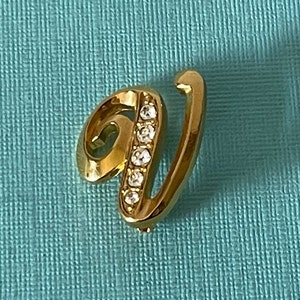 Peut inclure: Une broche en forme de lettre V dorée avec des strass transparents. La lettre est dans un style d'écriture cursive.