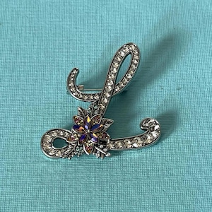 May include: Silver-toned brooch in the shape of the letter "L", encrusted with clear crystals. A flower with purple, pink, and gold petals adorns the base of the letter. The brooch is set against a light blue background.