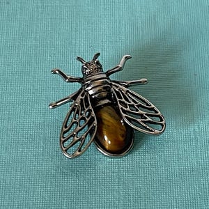 May include: A silver bee brooch with a tiger's eye gemstone. The bee's wings are detailed and intricate.