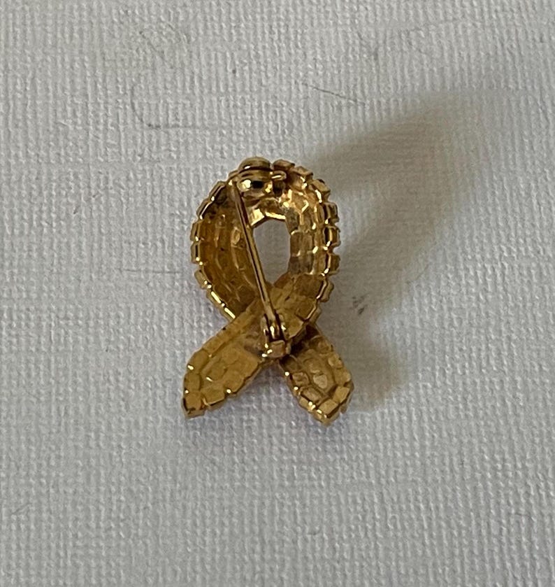 May include: A gold-toned ribbon brooch with a pin on the back. The ribbon is made of small, raised squares.
