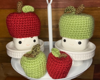 May include: A white tiered serving stand with four crocheted apple-shaped marshmallows. Two of the marshmallows are green and two are red.