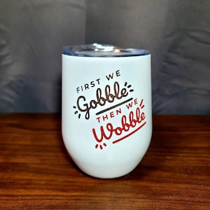 Puede incluir: Un vaso blanco de acero inoxidable con un gráfico rojo y negro que dice "First we Gobble then we Wobble".