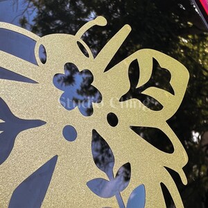May include: A close-up of a gold butterfly-shaped decal with detailed cut-out patterns. The decal is applied to a black surface, with the text "Studio Chings" visible. The design incorporates floral and leaf motifs, creating a decorative element.