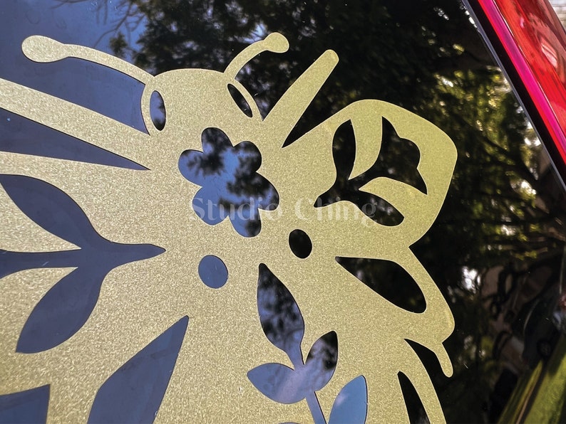 May include: A close-up of a gold butterfly-shaped decal with detailed cut-out patterns. The decal is applied to a black surface, with the text "Studio Chings" visible. The design incorporates floral and leaf motifs, creating a decorative element.