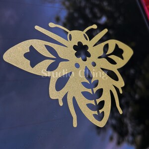 May include: A gold-coloured bee-shaped decal featuring floral and leaf cutouts. The decal is attached to a dark surface, possibly a car window. The text "Studio Ching" is visible on the decal.