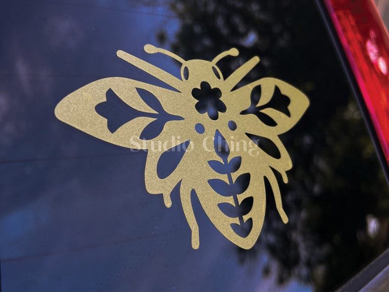 May include: A gold-coloured bee-shaped decal featuring floral and leaf cutouts. The decal is attached to a dark surface, possibly a car window. The text "Studio Ching" is visible on the decal.