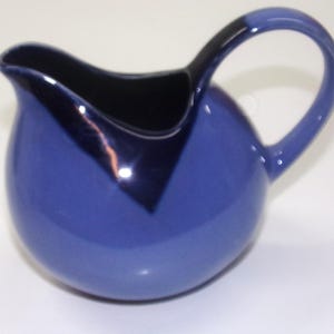 May include: A blue ceramic pitcher with a rounded body and a curved handle. The interior and a section of the rim are black. The pitcher has a glossy finish and is designed for pouring liquids.