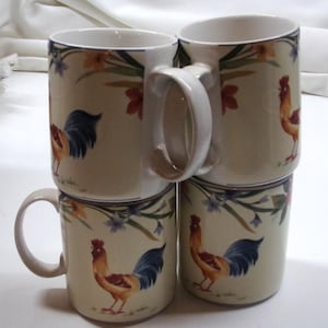 May include: Four ceramic mugs stacked, featuring a rooster design. The mugs have a cream-colored base with a floral border and a rooster illustration in shades of brown, blue, and yellow. Each mug has a white handle.