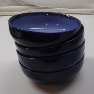 May include: A stack of four navy blue ceramic bowls. The bowls have a glossy finish and a simple, modern design. The interior of each bowl is a lighter shade of blue, creating a contrast with the darker exterior.