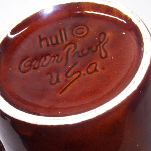 May include: Close-up of a brown ceramic mug with a white rim. The bottom of the mug features the embossed text "hull", "Oven Proof", and "U.S.A.". The mug has a glossy finish and a dark brown color.