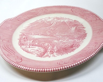 Könnte beinhalten: Eine ovale Servierplatte aus Keramik mit einem rosa-weißen Transferdruckmuster. Die Mitte zeigt eine Landschaft mit Bergen, einem See und Figuren, während der breite Rand ein dekoratives Schnörkelmuster und einen gestreiften Rand aufweist.