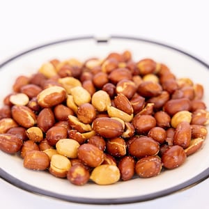 May include: A white plate with a black rim holds a pile of roasted peanuts. The peanuts are a mix of light brown and dark brown.