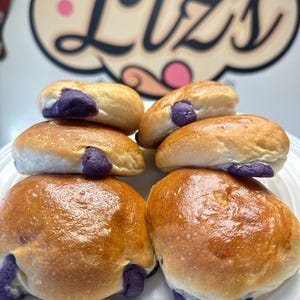 May include: Six golden brown ube bread rolls with purple ube filling on a white plate.