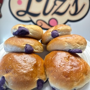 May include: Six golden brown ube bread rolls with purple ube filling on a white plate.