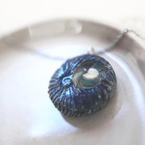 Pode incluir: Um colar azul e prateado com um pingente em forma de concha. O pingente tem um centro de vidro transparente.