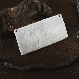 May include: A rectangular white pendant with a textured surface hangs from a copper chain. A vintage key and a piece of driftwood are also in the frame, set against a dark wooden background. The pendant is a neutral color.